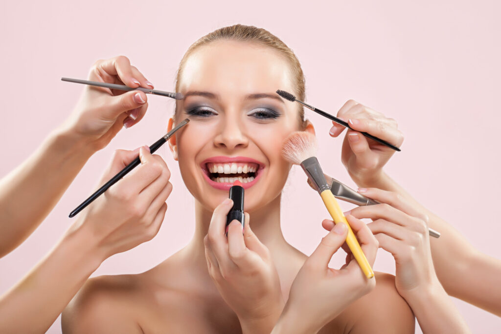 Frau wird gleichzeitig von mehreren Händen mit Make-up geschminkt und lacht