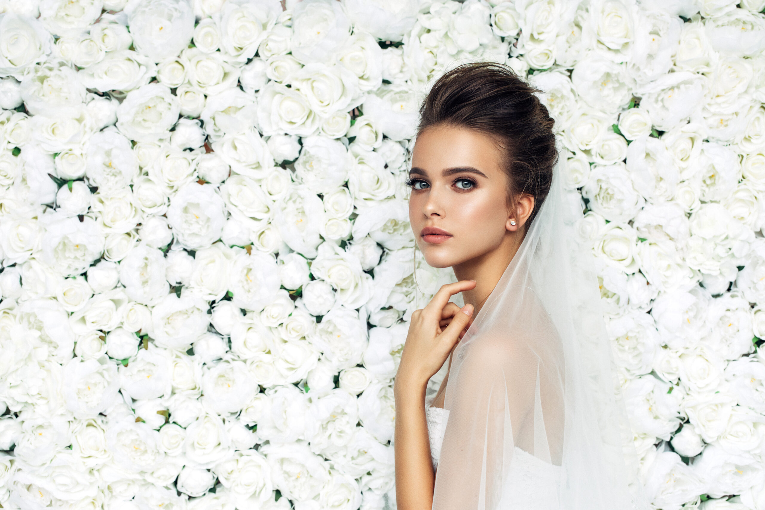 Elegante Braut mit Hochsteckfrisur und Schleier vor Blumenwand
